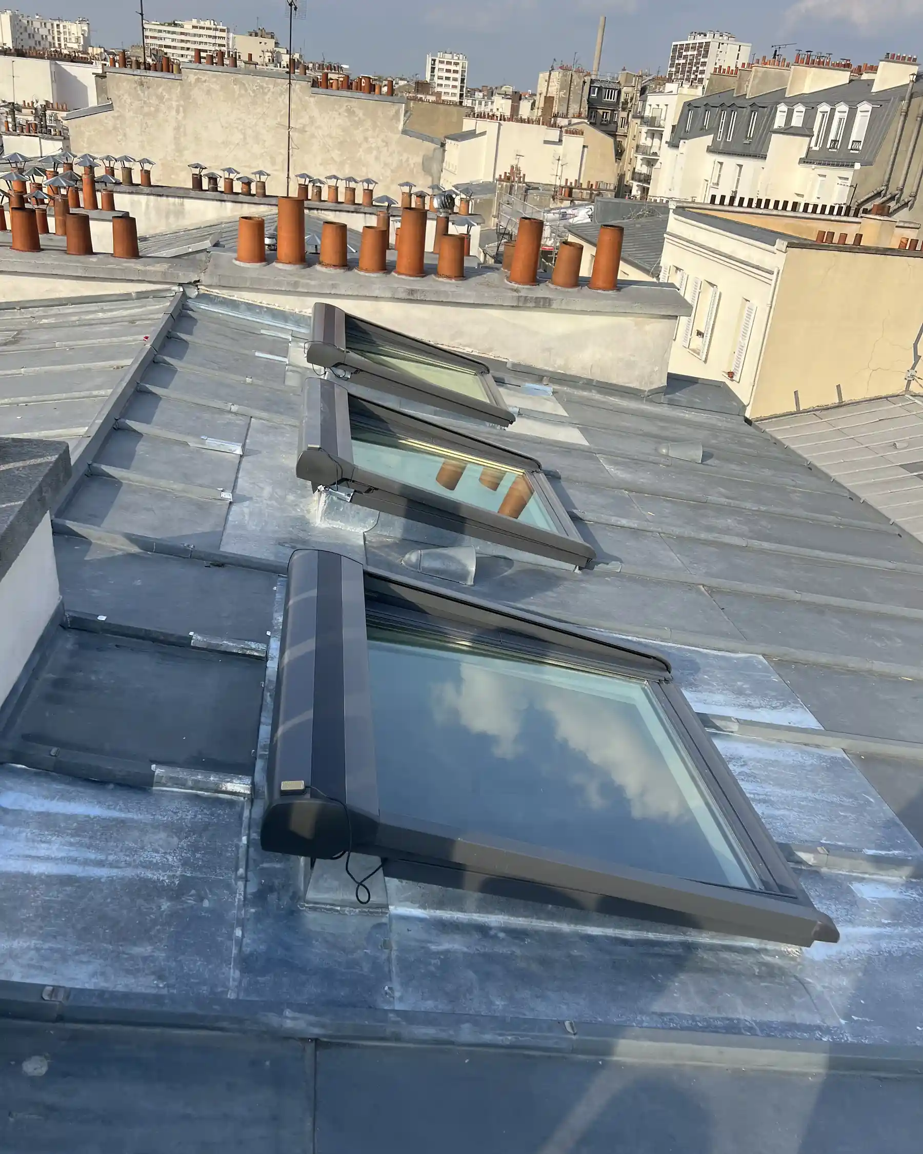 Trois fenêtres de toit VELUX posées sur une couverture zinc, cheminées et toits en arrière-plan